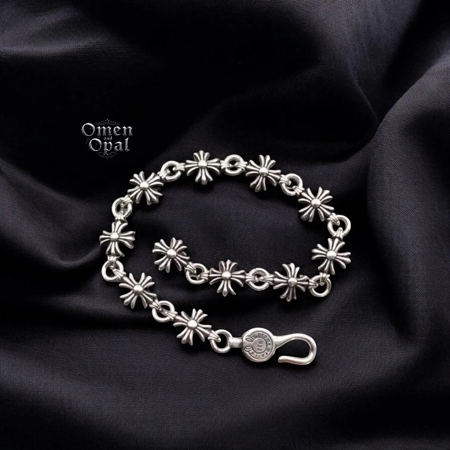 A silver-tone chain charm bracelet with a gothic cross, heart, and dice charms displayed on a black fabric background.