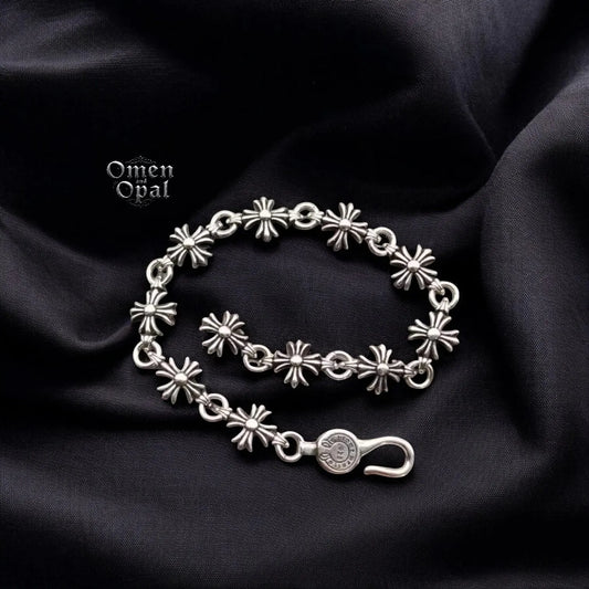 A silver-tone chain charm bracelet with a gothic cross, heart, and dice charms displayed on a black fabric background.